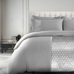 Elegant bedroom with a plush, silver comforter featuring a geometric pattern, matching pillows, and a tufted headboard. Minimalist decor.