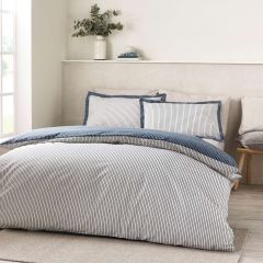 Bright, serene bedroom featuring a neatly made bed with navy-and-white pinstripe duvet and pillows, plants, books, and soft natural light.