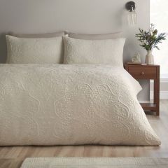 Cream paisley quilt and matching pillows adorn a neatly made bed beside a wooden nightstand with flowers, creating a calm, elegant bedroom.