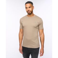 A man with short hair and a beard is wearing a fitted taupe T-shirt and black pants. He stands against a plain white background with a relaxed expression.