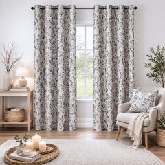 Cozy living room with leaf-patterned curtains on a window, a gray armchair with a matching pillow and blanket, and a wooden table with a lamp. Candles on a woven tray and a rug add warmth.