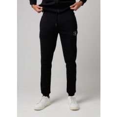 A person stands wearing black jogger pants, pulling at a black zipper hoodie. They sport white sneakers on a plain gray background.