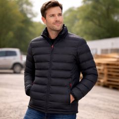 Man wearing a black padded work jacket outdoors, shown standing with hands in pockets on a worksite background.