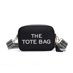 Black rectangular tote bag with white text "THE TOTE BAG" on the front. Features a black-and-white patterned strap and a black tassel accent.