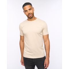 A man with short hair and a beard is wearing a fitted sand T-shirt and black pants. He stands against a plain white background with a relaxed expression.