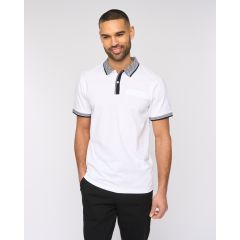 Man smiling in a white polo shirt with black trim, standing against a plain background. His relaxed pose conveys a casual, cheerful vibe.