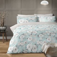 A cozy bedroom with a floral-themed duvet cover in pastel blue and white. A white wall lamp hangs above, and a potted plant sits on a wooden floor.