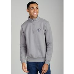 A smiling man in a gray quarter-zip sweater with a blue logo stands against a plain background. He's wearing blue jeans, conveying a casual, happy tone.