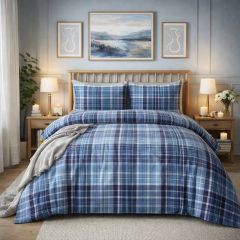 A cozy bedroom with a blue plaid bedspread, wooden headboard, bedside tables with lamps, candles, and framed art on a light blue wall.