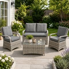 Modern grey rattan garden furniture set featuring a two-seater sofa, two armchairs and a glass-top coffee table arranged on a landscaped patio in a sunny back garden.
