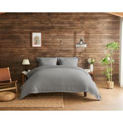 Inviting bedroom with grey bedding on a wooden bed, against a rustic wood-paneled wall. Decor includes a woven chair, plants, and soft lighting.