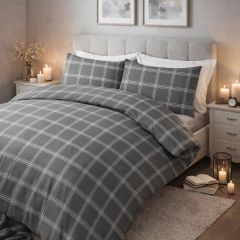 A neatly made bed with a gray plaid duvet and pillows in a bright room with a large window and light wooden floor.