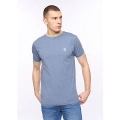 Man in a blue T-shirt with a white logo, looking to his left. He wears jeans, stands with hands in pockets against a plain white background.