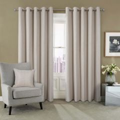 Beige curtains hang beside a window with a city view. A gray armchair with a matching pillow is on the left. A framed floral art piece and a vase with white lilies are on a metallic side table to the right. The tone is calm and modern.