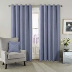 Blue curtains hang beside a window with a city view. A gray armchair with a matching pillow is on the left. A framed floral art piece and a vase with white lilies are on a metallic side table to the right. The tone is calm and modern.