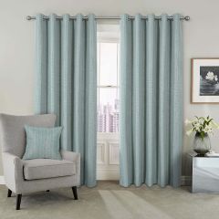 Green curtains hang beside a window with a city view. A gray armchair with a matching pillow is on the left. A framed floral art piece and a vase with white lilies are on a metallic side table to the right. The tone is calm and modern.