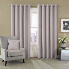 Taupe curtains hang beside a window with a city view. A gray armchair with a matching pillow is on the left. A framed floral art piece and a vase with white lilies are on a metallic side table to the right. The tone is calm and modern.