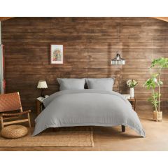 Inviting bedroom with grey bedding on a wooden bed, against a rustic wood-paneled wall. Decor includes a woven chair, plants, and soft lighting.