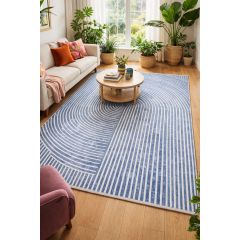 A cozy living room with a large blue and white geometric rug, a beige sofa with colorful cushions, and potted plants by a bright window. The mood is calm and inviting.