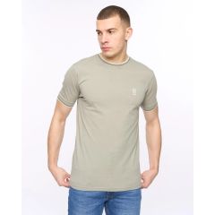 Young man wearing a fitted light sage green T-shirt with a small logo on the chest, paired with blue jeans. He gazes to the side against a plain white background, conveying a relaxed and confident vibe.