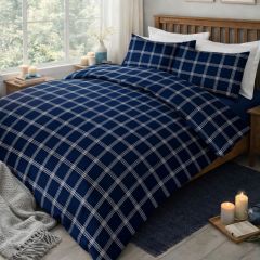 A neatly made bed with a navy blue and white checked duvet set in a bright room with a window, a vase, and small decor items.