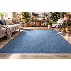 A cozy living room with a large blue rug, a gray sofa, and armchair adorned with pillows. Plants and warm lighting create a serene, inviting atmosphere.