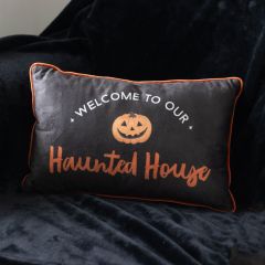 Black pillow with orange trim, featuring a pumpkin and the text "Welcome to Our Haunted House," placed on a dark fabric background.