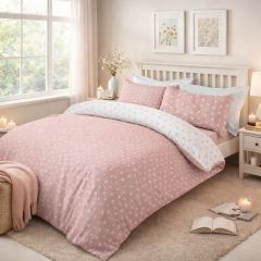 A bed with a white wooden frame, featuring pink bedding with small white floral patterns. Two matching pillows and a window with greenery outside.