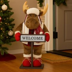 Plush Christmas moose decoration holding a WELCOME sign, standing inside a festive doorway with garlands and lights