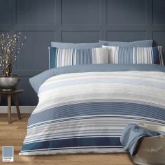 A cozy bedroom features a bed with Portsmouth striped bedding in shades of blue and white against a paneled blue wall. A side table holds a plant, creating a serene atmosphere.