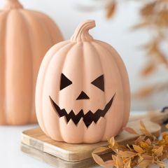 Ceramic pumpkin with a carved, mischievous face on a wooden board, surrounded by autumn leaves.