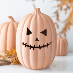 A ceramic pumpkin with a carved jack-o'-lantern face sits on a white surface, surrounded by autumn leaves and smaller pumpkins.