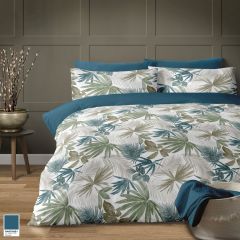 A cozy bedroom setting with a bed featuring a tropical Ripley Fern leaf-patterned duvet in muted greens and blues. A pot with tall branches sits on a wooden floor.