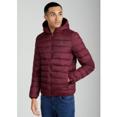 A person in a burgundy puffer jacket with a hood stands against a plain gray background. They have one hand in a pocket, wearing blue jeans, looking off-camera.