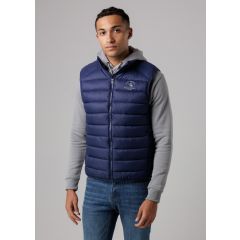 A man wearing a navy quilted vest over a gray hoodie and blue jeans stands against a plain background. He looks confidently at the camera.