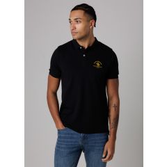 Man in a black polo shirt with a yellow logo, standing against a neutral background. He has tattoos on his arm and wears blue jeans, looking to the side.