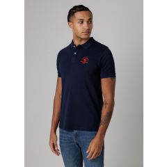 Young man in a navy polo with red logo, looking to the side. He wears blue jeans and has tattoos on his arm, standing against a gray background.