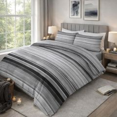 A neatly made bed with a gray striped duvet and pillows, in a bright room with a window, wooden floor, and small decor items.